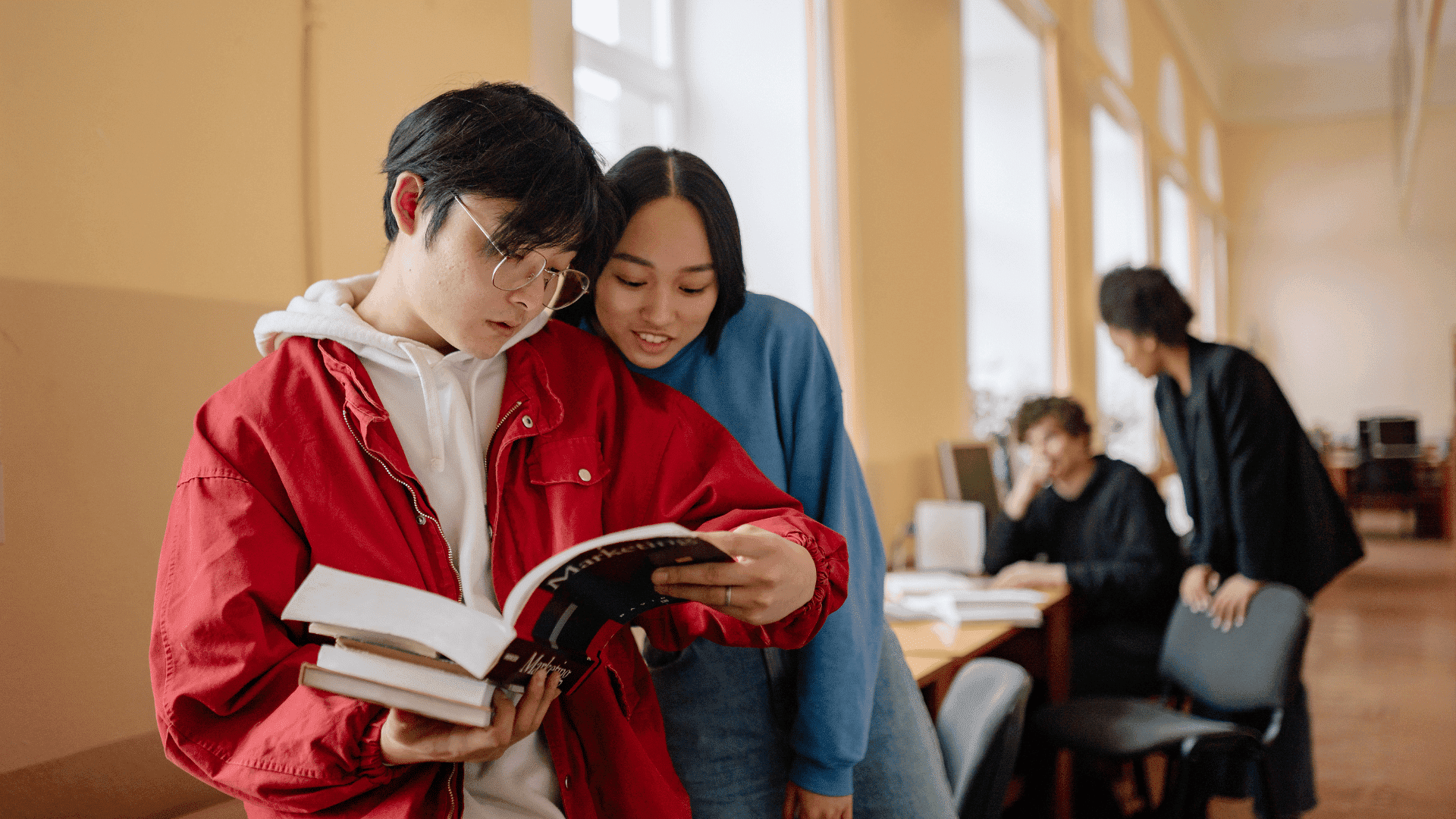 Students reading a book together in a classroom, with more students working in the background.